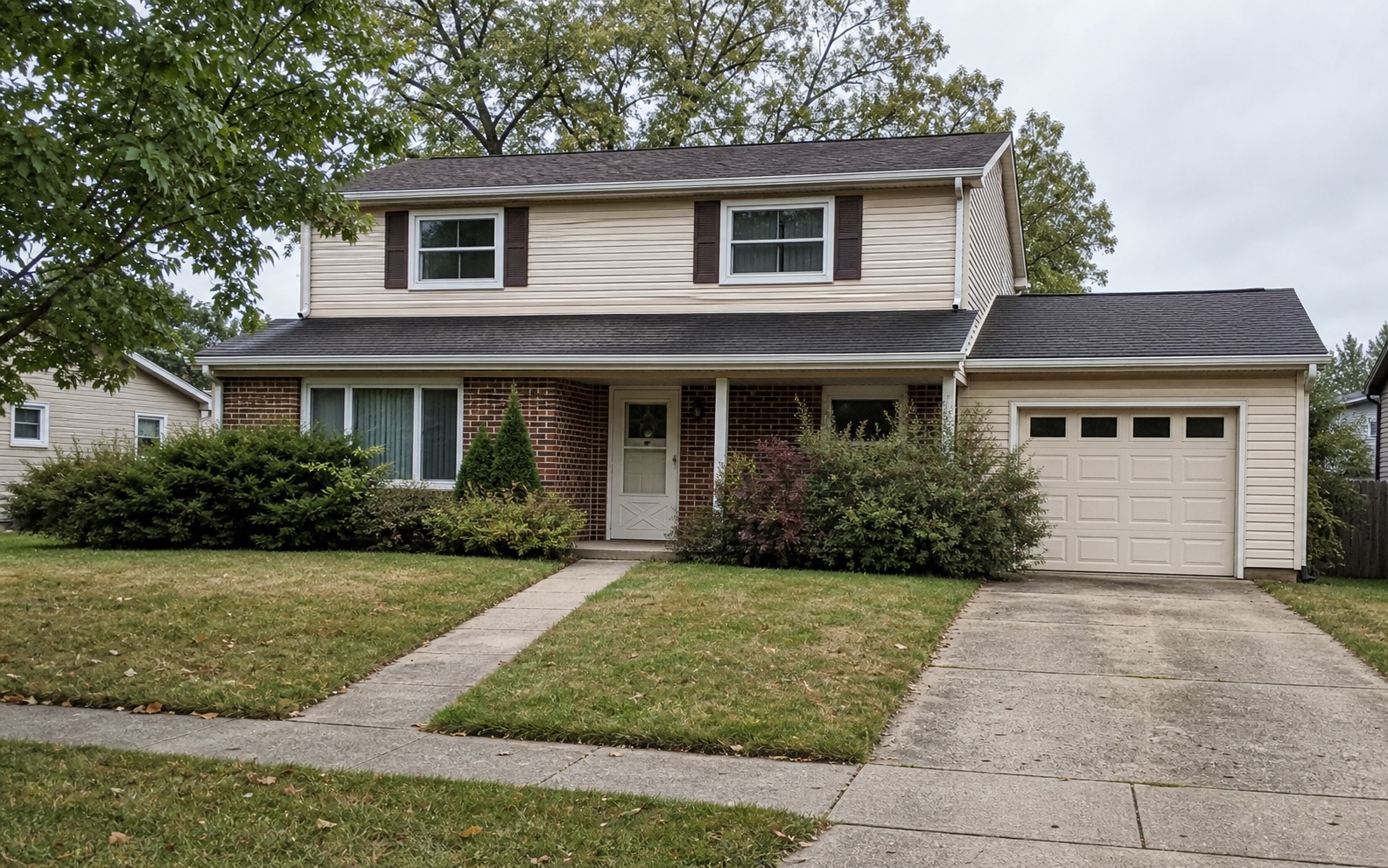 Before photo of a dated suburban house exterior with beige siding, brick lower facade, one-story garage wing, covered front porch, concrete walkway, driveway, patchy lawn, and overgrown shrubs.