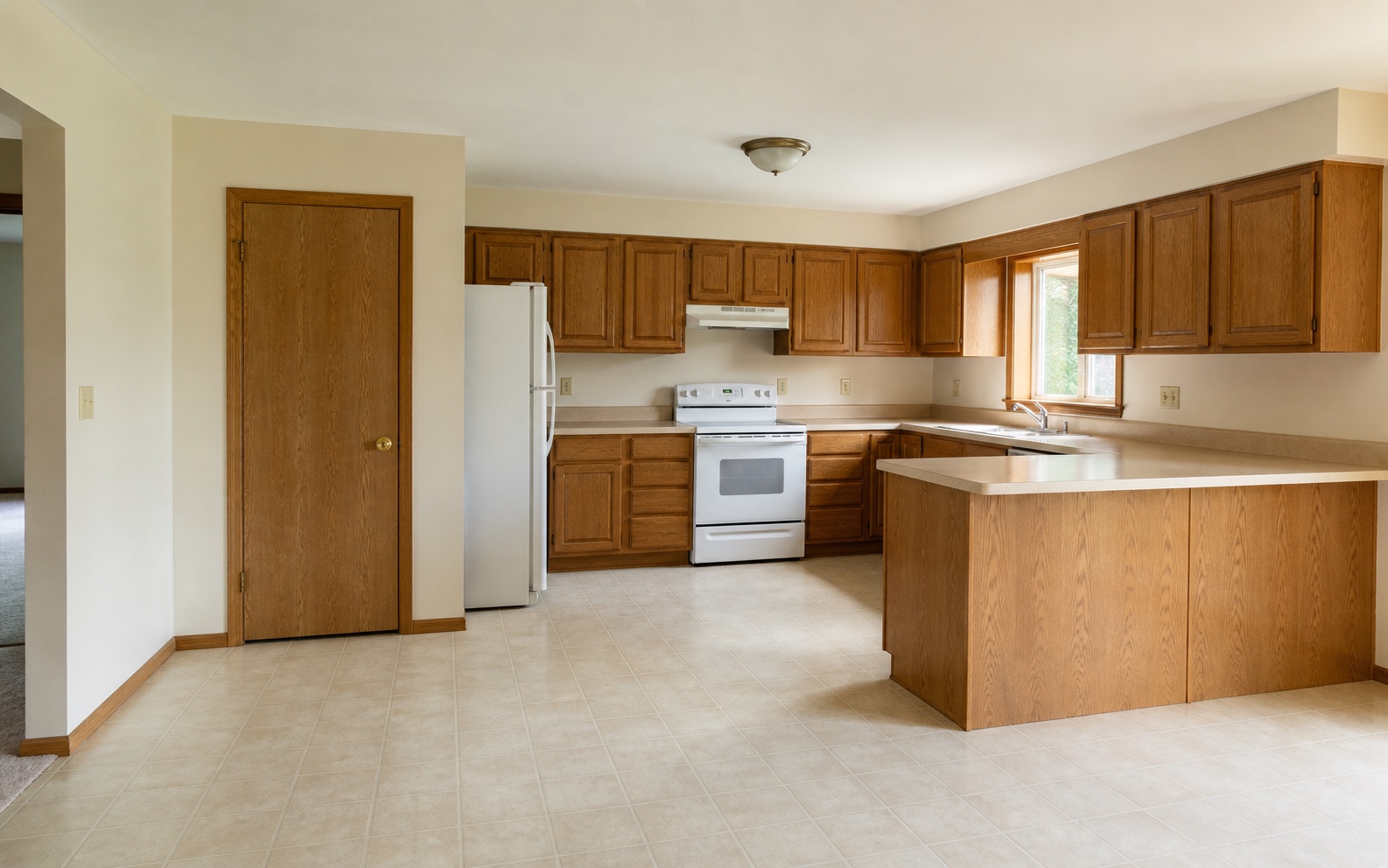 Before photo of a dated honey-oak kitchen with the original door, window, cabinet runs, appliances, and peninsula visible.