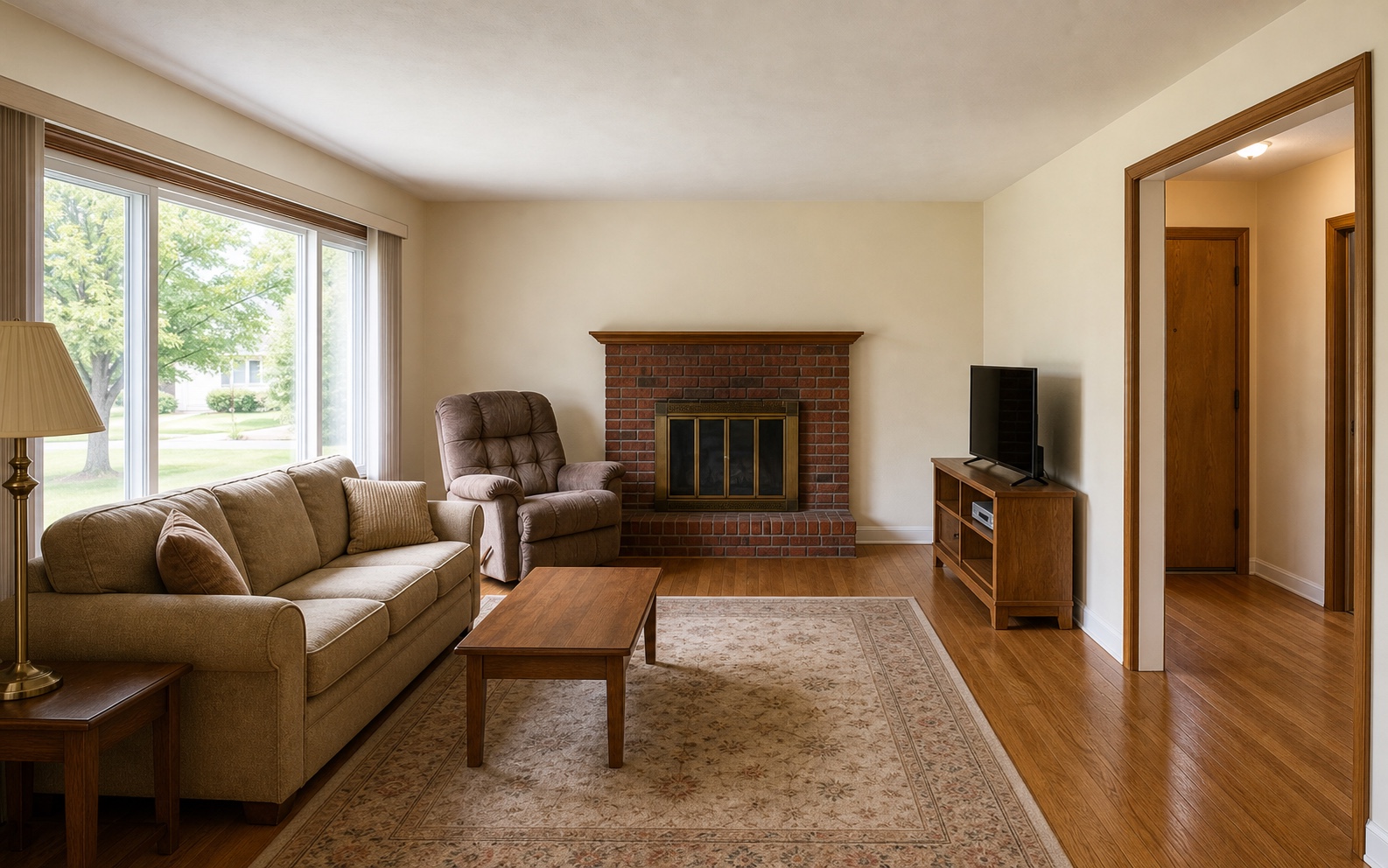 Before photo of a dated living room with vertical blinds, bulky seating, brick fireplace, brass fireplace doors, older wood TV console, beige walls, wood trim, and hardwood floor.
